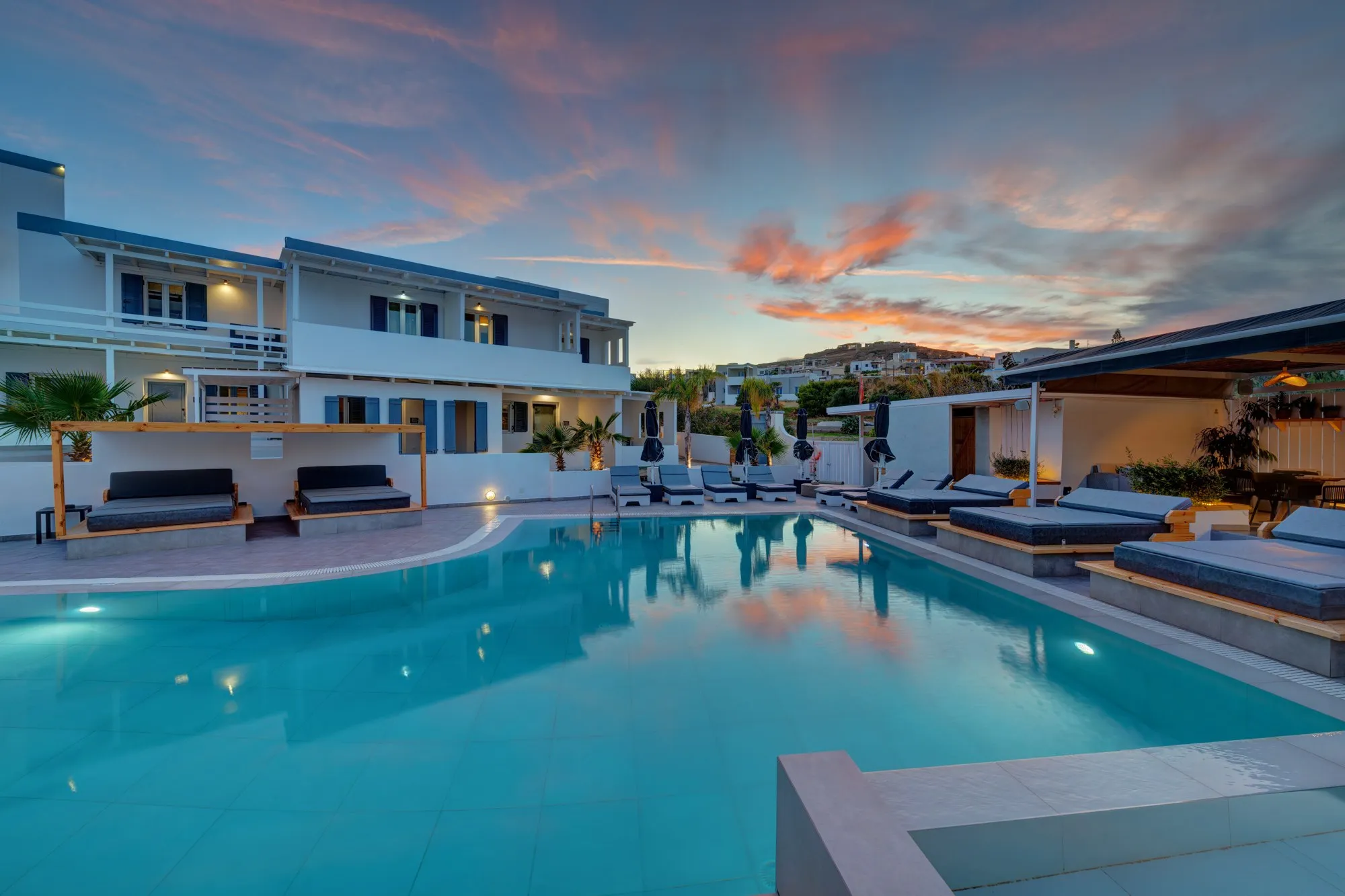 The pool at dusk under a pink sky, with daybeds along the water and the white buildings of the club behind