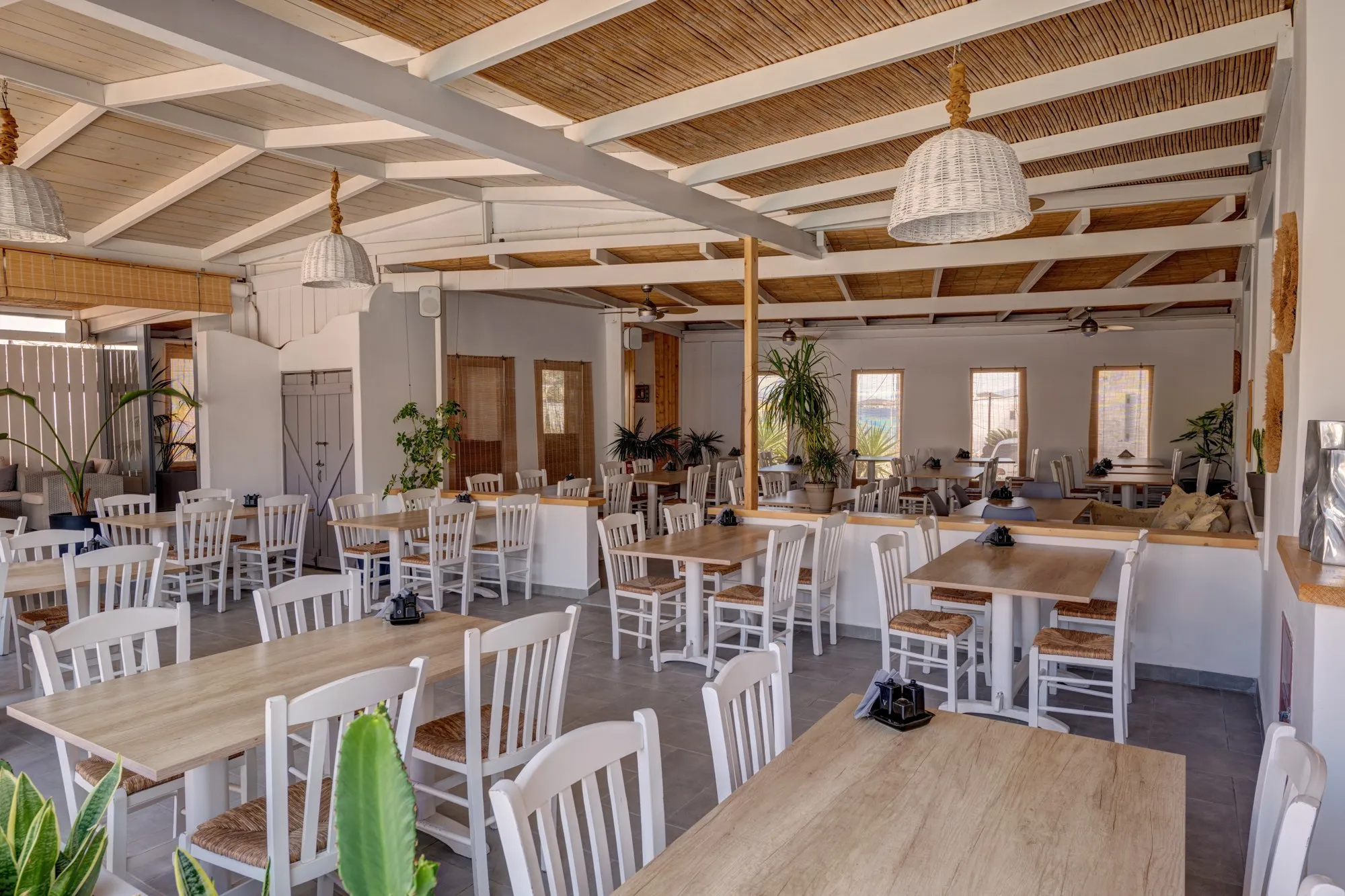 The restaurant under its reed roof, with white wooden chairs, wicker lamps and plants